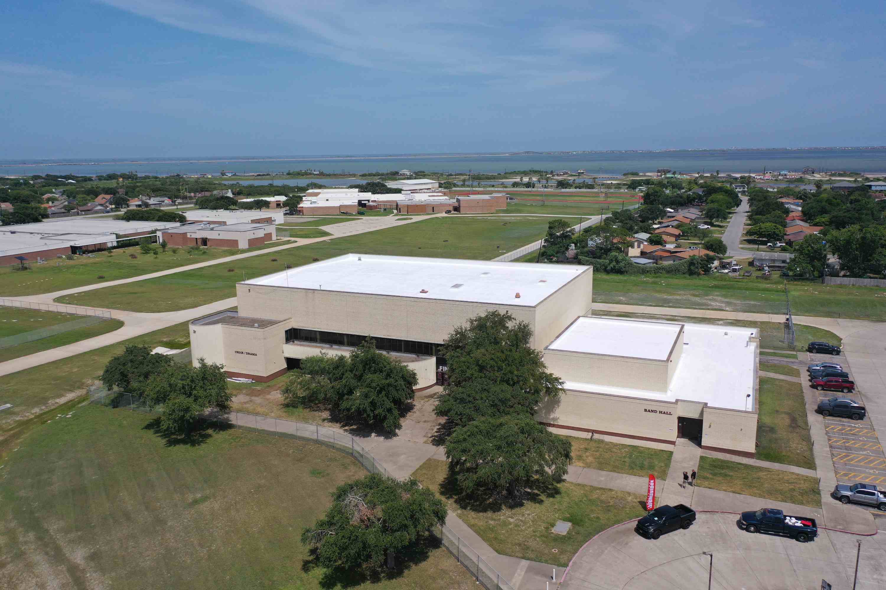 Aerial view of a large school building with a white roof, surrounded by trees, parked cars, grassy fields, and nearby residential areas, with a body of water in the background.