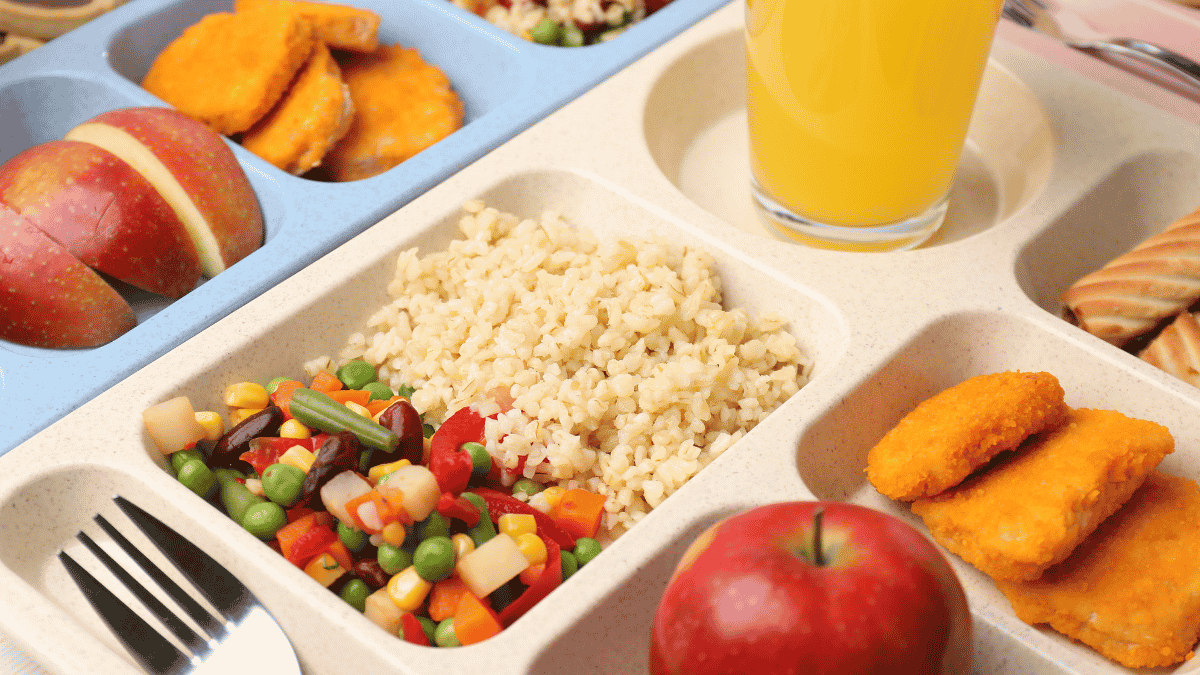 A lunch tray with rice, mixed vegetables, apple slices, a whole apple, bread, orange juice, and breaded nuggets in separate compartments.